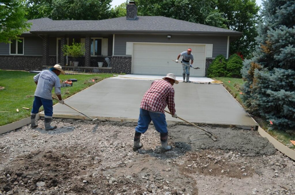 Crew raking and finishing a freshly poured concrete driveway in front of a house